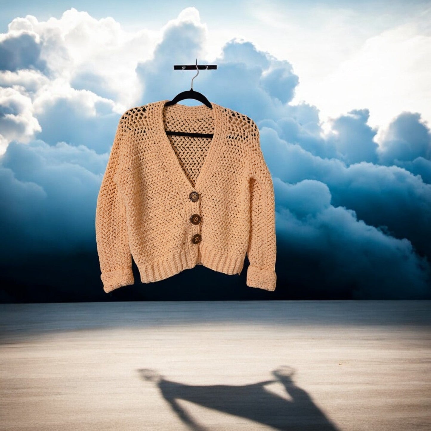 Handmade peach cardigan sweater, cropped with button detail, on hanger against cloudy sky.