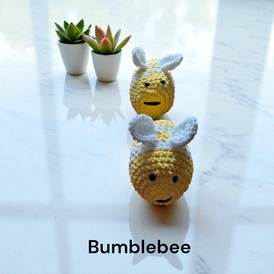 handcrafted yellow and white amigurumi bumblebee plush on reflective surface with small potted plants in background