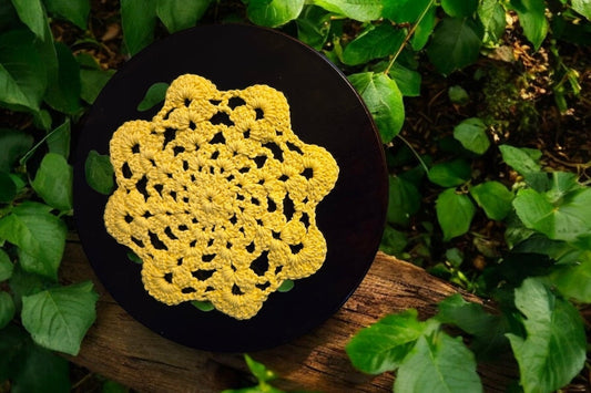 Vintage yellow crochet doily made from soft cotton yarn on dark wooden surface surrounded by green leaves