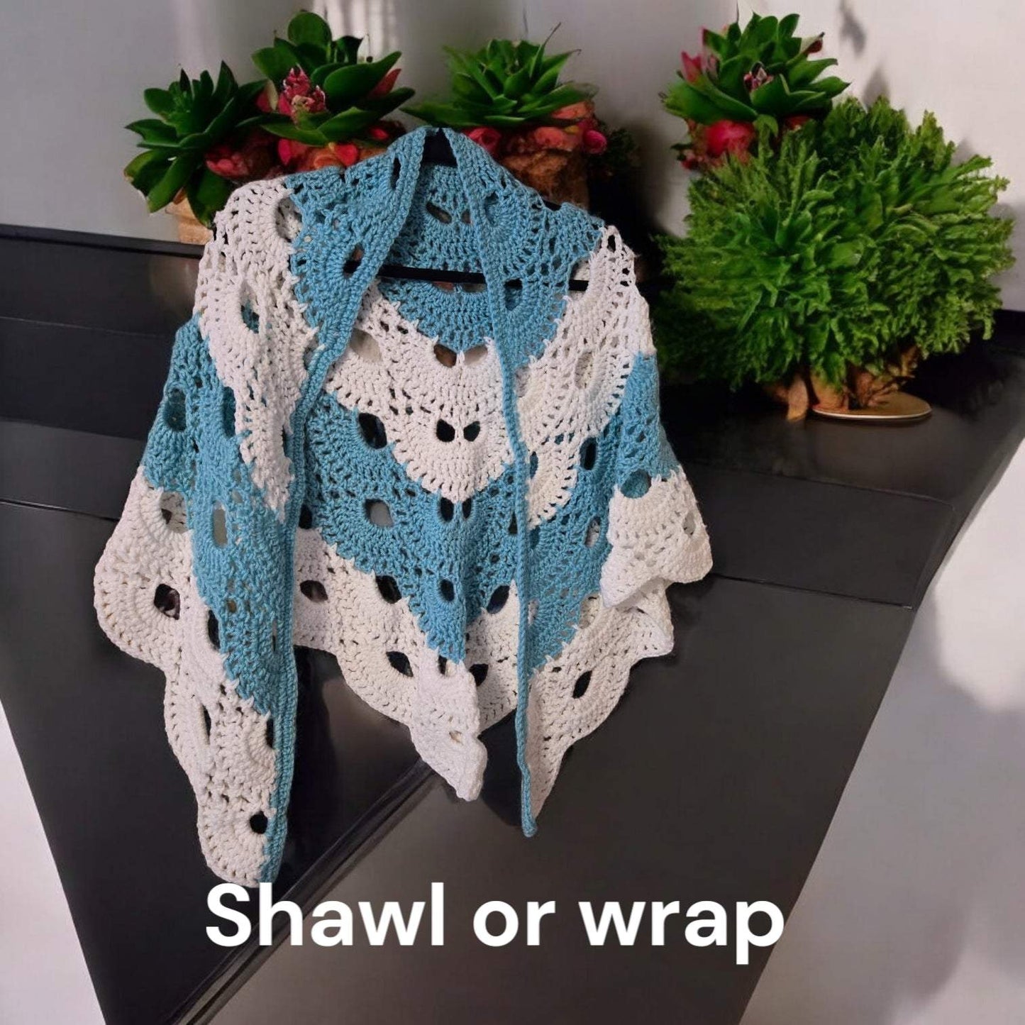 Designer handmade crocheted shawl in blue and white cotton yarn displayed on a black surface with plants in the background